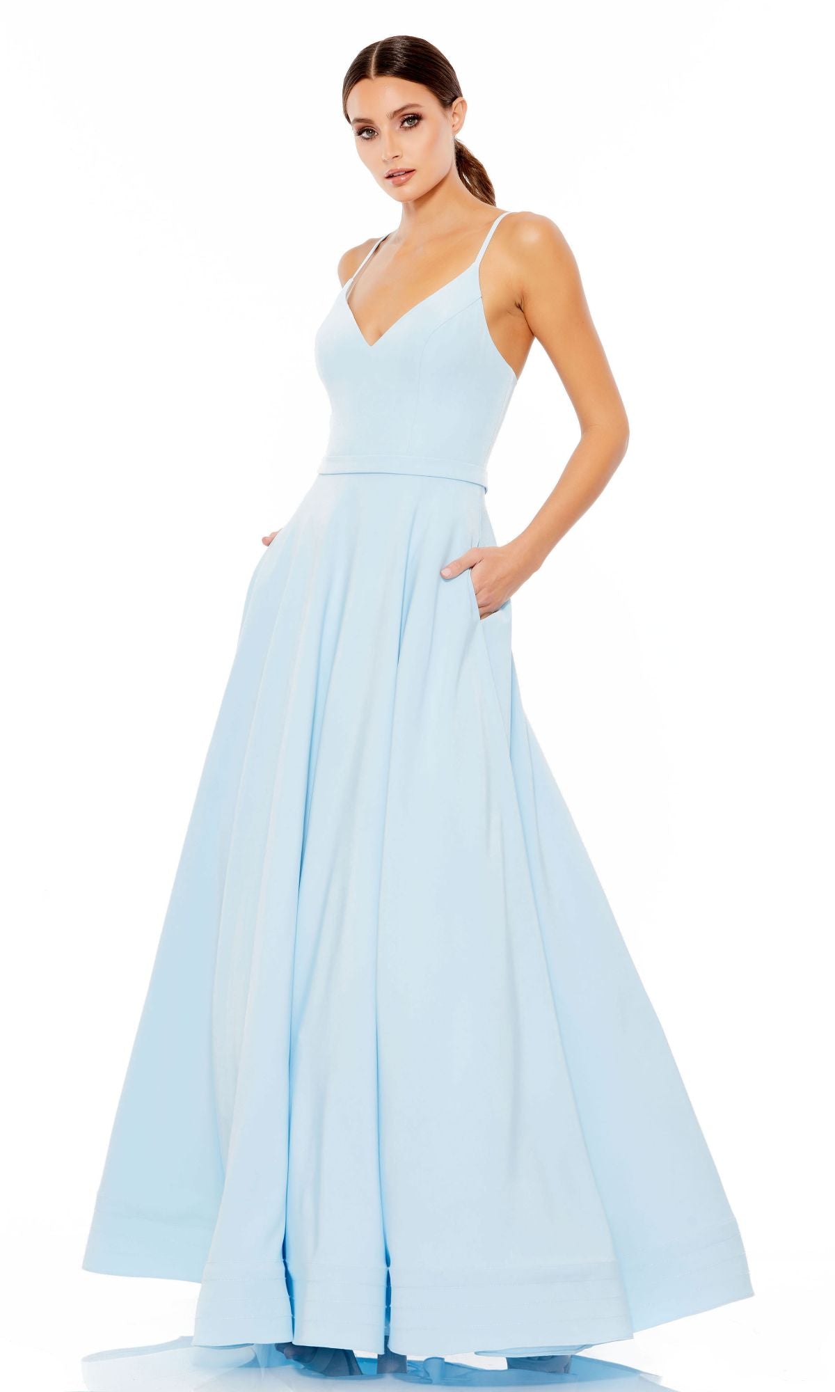 A woman stands against a white background wearing the Classic Long A-Line Formal Dress: Mac Duggal 48855, a sleeveless, light blue floor-length gown featuring a V-neckline and side pockets.